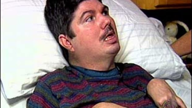 Terry Wallis lying in a hospital bed being interviewed after waking up from his 19 year coma wearing a striped shirt.