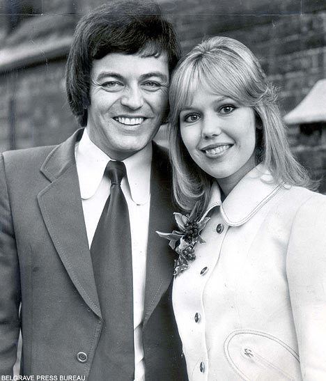 Tony Blackburn and Tessa Wyatt are smiling on their wedding day at Caxton Hall, London, on 2nd March 1972. Tony is wearing a long sleeve under a necktie and a coat while Tessa is wearing a coat with a flower on the left side