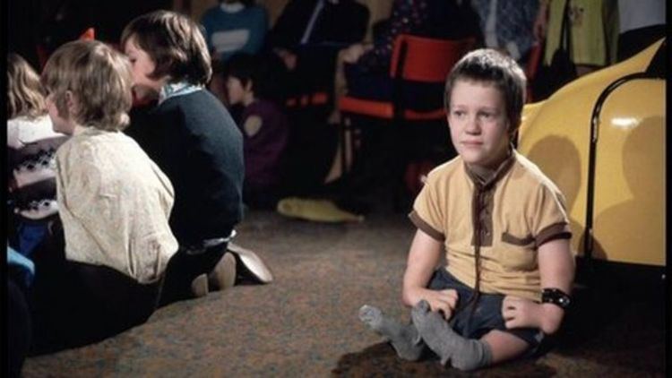 A Thalidomide victim sitting while looking at something together with other victims who had battled for years to get justice in the background. He has brown hair, a wrist band on his right, gray socks, wearing a yellow and brown mid-buttoned shirt and a short.