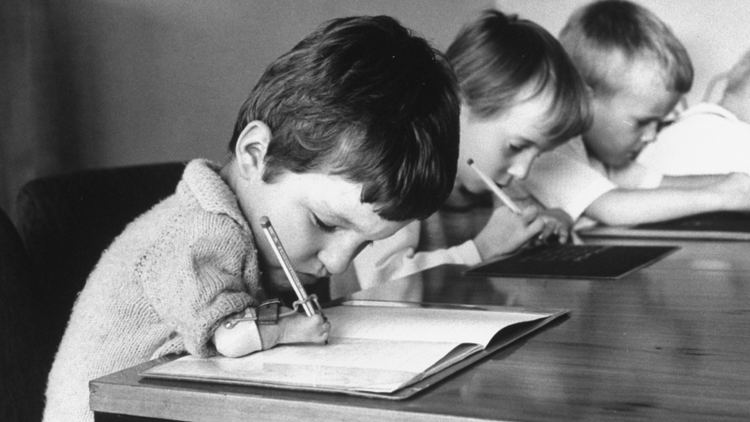 The child in the front having difficulty in writing due to his malformed hands affected by Thalidomide along with two other children. In the front, the child is sitting and holding a pencil clipped on the knitted polo he's wearing and is writing on a piece of paper placed on the table. The child next to him is also sitting and holding a pencil, wearing a long sleeves shirt while the other is sitting and leaning his right elbow on the table wearing a polo shirt.