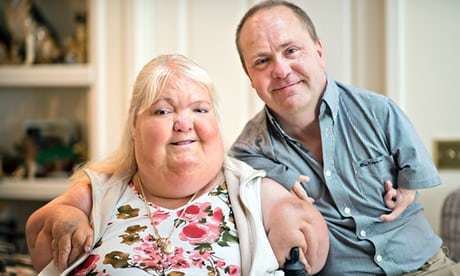 Louise Medus-Mansell smiling with her husband, Darren who suffered from body malformations due to Thalidomide. Louise with a blonde hair, wearing a floral dress and a necklace while Darren wearing a blue shirt