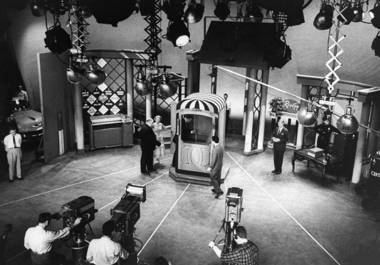 In the set of “The $64,000 Question” The isolation booth is at the center, it has lights and cameras with the hosts, a man on the left is serious, hands behind wearing white long sleeves, gray pants, and black shoes. 2nd from left are two hosts, a man (left) is turning his back, bald, wearing a black suit, black pants, and black shoes. 3rd from the left is a woman, wearing a white dress. On right is a man standing, wearing white long sleeves under a black suit, black pants, and black shoes. On the bottom left, a man is holding a camera with his right hand, has black hair, and wears white long sleeves and black pants. 2nd from the bottom left is a man also holding the camera, has black hair, and wears white long sleeves.