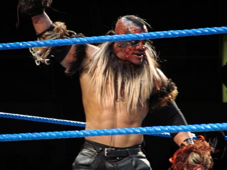 Martin Wright topless, wearing black pants, and a black belt with hair on his arms and neck inside a wrestling ring.