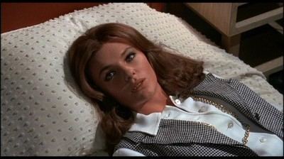 Jacqueline Bisset lying on the bed with blonde hair in a scene from the 1969 film "The First Time" and wearing a white long sleeve blouse and black and white checkered vest and pearl necklace