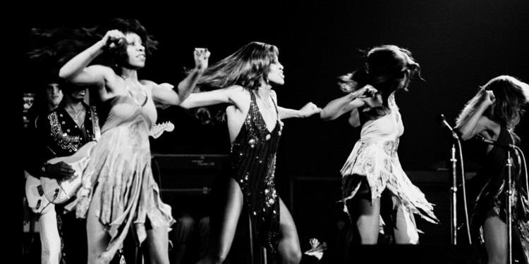 In black and white on a stage with mic and instruments the Ikettes is Dancing, from left, a man standing playing a guitar has black hair wearing a black shirt and white pants, 2nd from left, a woman is serious dancing, with her hands up has black hair wearing a white dress, 3rd from left, a woman is serious, dancing, hands up, has black hair wearing a black shiny bathing suit, 4th from left, a woman is looking at her left, dancing, hands up, has black hair wearing a white dress, at the right is a woman dancing looking down, hands up has black hair wearing a black dress.