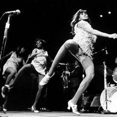 In black and white a group of woman the Ikettes dancing on a stage with mic stand and drums at the back, from left a woman is serious, dancing, looking down at her left, standing with her left foot and right foot up has black hair wearing a white dress and white shoes, in the middle a woman is smiling, looking at her right, standing with her left foot and right foot up has black hair wearing a white dress and white shoes, at the right is a woman is serious, looking up, dancing, standing with her left foot and right foot up has black hair wearing a  white panty under a white dress and white shoes.