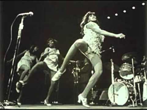 In black and white a group of woman the Ikettes dancing on a stage with mic stand and drums at the back, from left a woman is serious, dancing, looking down at her left, standing with her left foot and right foot up has black hair wearing a white dress and white shoes, in the middle a woman is smiling, looking at her right, standing with her left foot and right foot up has black hair wearing a white dress and white shoes, at the right is a woman is serious, looking up, dancing, standing with her left foot and right foot up has black hair wearing a  white panty under a white dress white dress and white shoes.