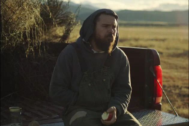 Clayne Crawford looking afar while sitting at the cargo bed and wearing a hoodie and jumpsuit in a scene from the 2020 drama film, The Killing of Two Lovers