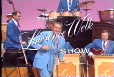 Lawrence Welk is smiling with a microphone in front, has black hair, wearing white long sleeves and a blue necktie under a blue suit, his right hand up, and left hand down. Behind is a man wearing white long sleeves and a blue necktie under a blue suit with drums, a man (left) is serious while looking down, wearing white long sleeves and a blue necktie under a blue suit and blue pants, a man (right) is serious, playing the guitar with his right hand, has a lectern in front, wearing white long sleeves and a blue necktie under a blue suit.