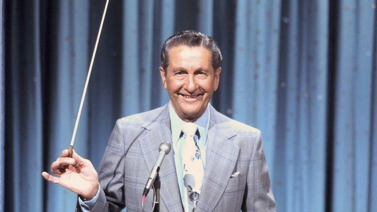 Lawrence Welk is smiling with a microphone in front, his right hand holding a stick. has black hair, wearing blue long sleeves and a light blue necktie with minimal design under a blue suit with lines.