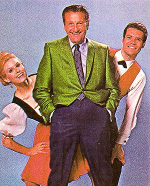 On left is Cissy smiling, has blonde hair, wearing a black and white top, an orange skirt, and her right hand holding her right leg. The middle is Lawrence Welk smiling, has black hair, wearing gray long sleeves, a  dark gray necktie with minimal design under a green suit, and dark blue pants, both hands inside his pocket. On (right) is Bobby Burgess smiling, has black hair,  his left hand holding his left leg, wearing a white long sleeve and an orange suit with a bow tie.