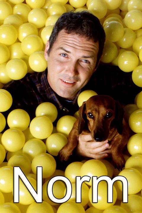 Norm MacDonald smiling and holding a dog while being surrounded by yellow balls and wearing checkered long sleeves