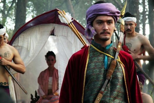 Ananda Everingham with a serious face, with an arrow and bow in a movie scene from The Outrage, a 2011 Thai Drama movie. He is wearing a purple cloth on his head, and a multi-colored robe while at the back, Chermarn Boonyasak sitting and wearing a pink and white dress.