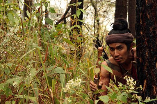 Dom Heatrakul with a serious face while hiding at the back of a tree with a forest in the background, holding a sword and wearing a brown cloth on his head in a movie scene from The Outrage, a 2011 Thai Drama movie. He has body tattoos and is topless.