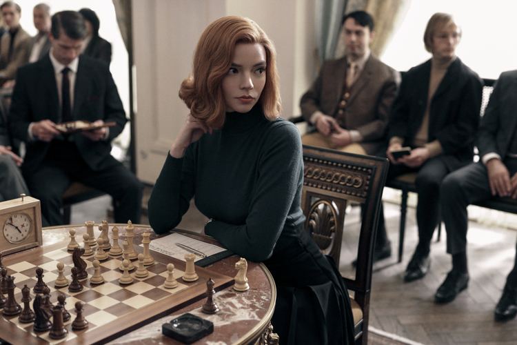 Anya Taylor-Joy with a serious face during a chess tournament and surrounded by other players, with short blonde hair, and wearing a black dress in a movie scene from The Queen'S Gambit (2020).