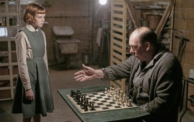 Isla Johnston as Young Beth Harmon looking at Bill Camp	as Mr. Shaibel playing chess. Isla wearing a gray dress over white long sleeves while Bill wearing a gray jacket in a movie scene from The Queen'S Gambit (2020).