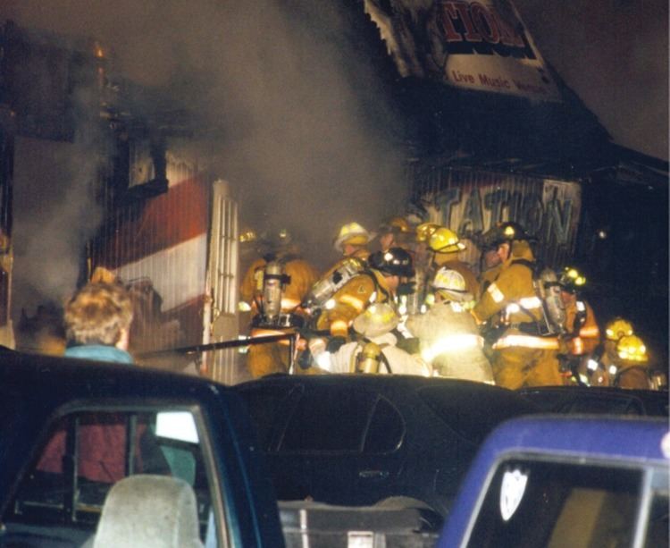 An image of The Station night club fire where metal handrail and parked vehicles impede recovery efforts at the front entryway by numbers of firefighters. And a window to the bar room is at left.
