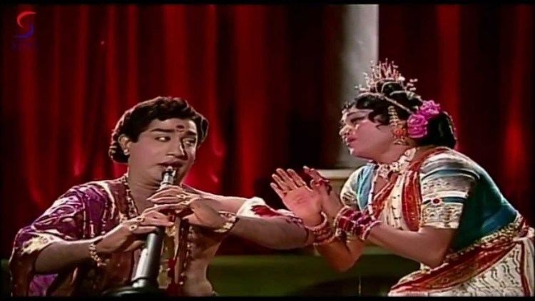 Shivaji Ganesan plays the flute while Padmini is singing while looking at each other. Shivaji wearing a maroon Indian shirt while Padmini wearing a maroon and green Indian dress with accessories in a movie scene from Thillana Mohanambal (1968 film).