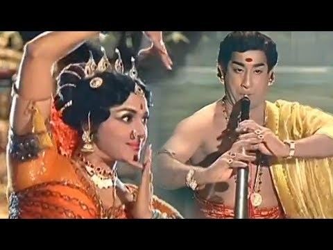 Padmini dancing while Shivaji Ganesan plays the flute. Padmini wearing an orange Indian dress with accessories while Shivaji is topless with a yellow scarf on his arm in a movie scene from Thillana Mohanambal (1968 film).