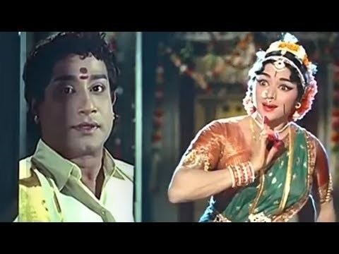 Shivaji Ganesan is smiling while Padmini with a surprised facial expression. Shivaji wearing a yellow polo shirt while Padmini wearing an Indian dress with accessories in a movie scene from Thillana Mohanambal (1968 film).