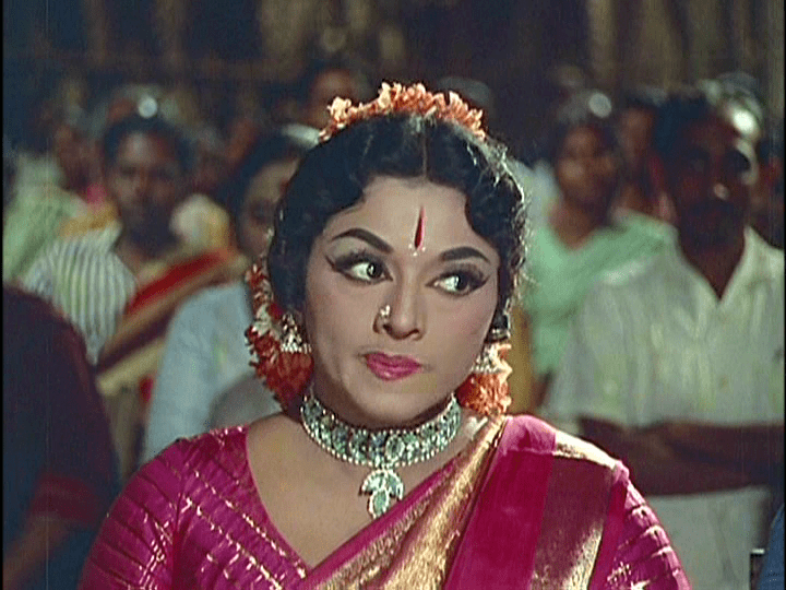 Padmini with a serious face, wearing a necklace, and a red Indian dress in a movie scene from Thillana Mohanambal (1968 film).
