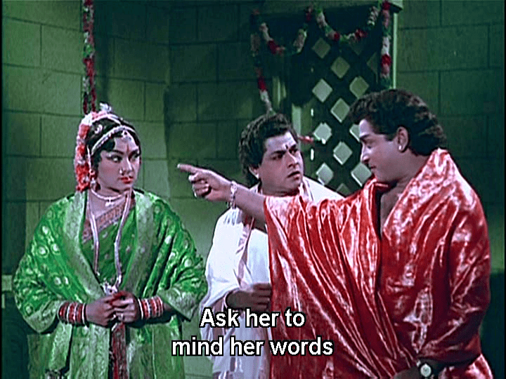 Padmini, T.S. Balaiah, and Shivaji Ganesan are talking at each other while Shivaji pointing his finger at Padmini. Padmini wearing a green Indian dress with accessories, T.S. wearing a white Indian suit, and Shivaji wearing a red robe in a movie scene from Thillana Mohanambal (1968 film).
