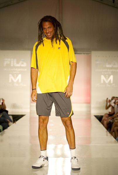 Thomas Blake smiling, with twist braids, and wearing a black and yellow t-shirt, gray shorts, white socks, and sneakers
