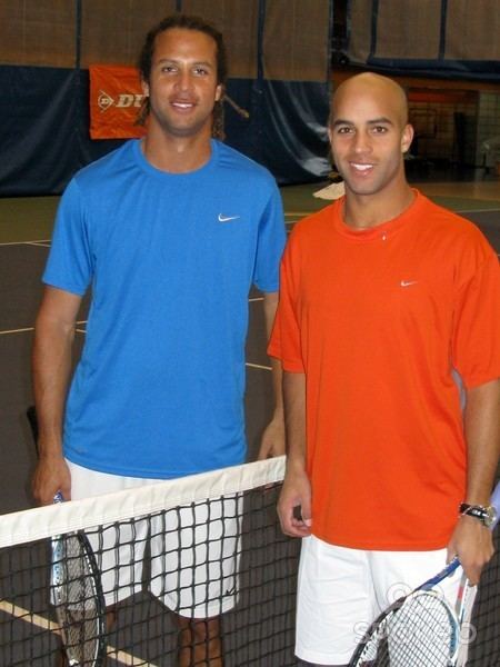 Thomas and James Blake are smiling and holding a racket. Thomas is wearing a blue shirt and white shorts while James is wearing an orange shirt