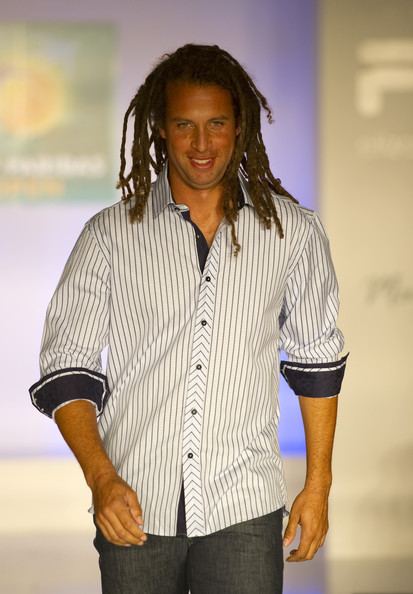 Thomas Blake smiling, with twist braids, and wearing a white and blue striped long sleeves and denim pants