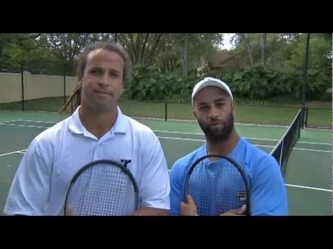 Thomas and James Blake are smiling and holding a racket. Thomas is wearing a white polo shirt while James is wearing a blue shirt and white cap