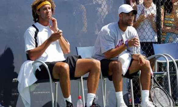 Thomas Blake and the man sitting beside him are looking afar while wearing a white t-shirt, black shorts, white socks, and rubber shoes