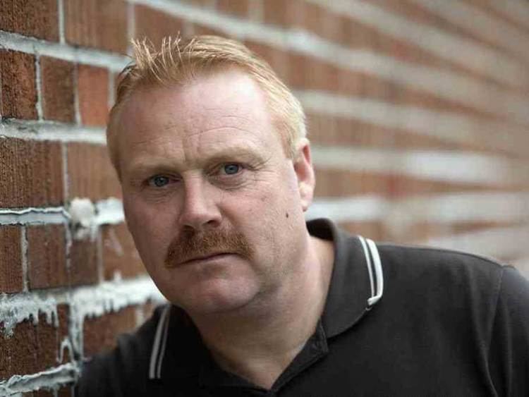 Thomas Craig posing near a wall with a bit of mustache and wearing a black polo shirt.