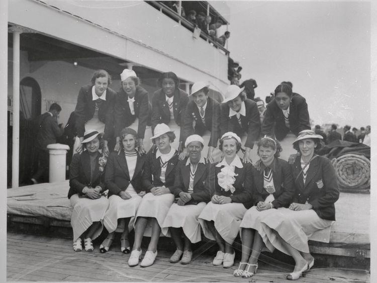 1936 US Women’s Olympic Track and Field Team are smiling, including Louise Stokes (back row, third from left) and Tidye Pickett (back row, far right). They are wearing hat, shoes, skirt, and blouse under a coat
