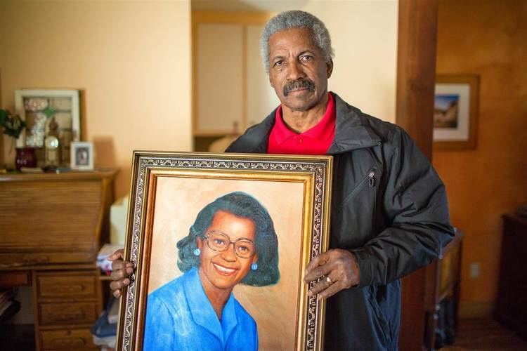 Tiequon Cox is serious, holding a painted portrait of a woman smiling, has black hair wearing eyeglasses, cyan earring and blue coat in a copper frame while standing in the middle of a room with a wooden cabinet at the back of his right arm and a doorway with painting on a brown wall at the back on his left, has gray old hair with black mustache wearing a red shirt under a black leather jacket with gold ring in his left hand.