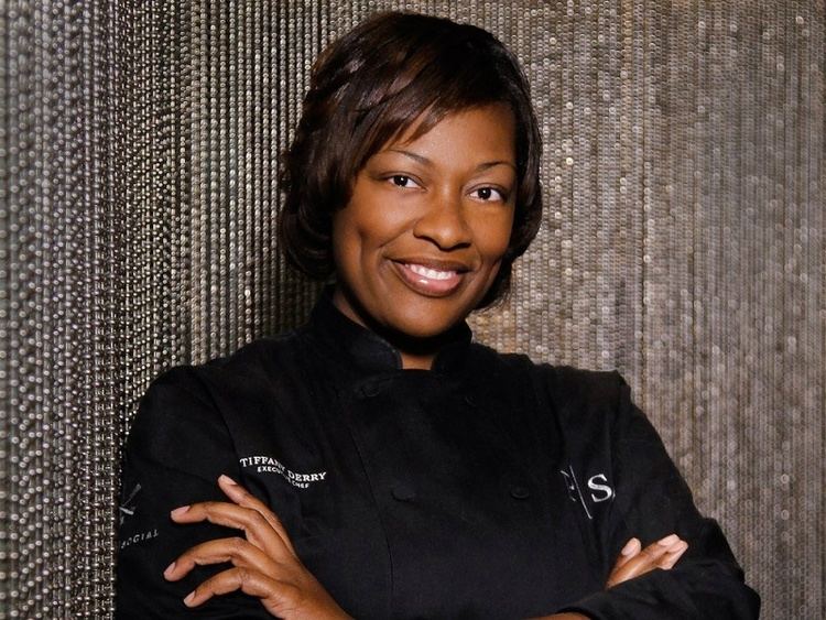 Tiffany Derry smiling with crossed arms on her chest, with short wavy hair, and wearing a black chef gown with white prints.
