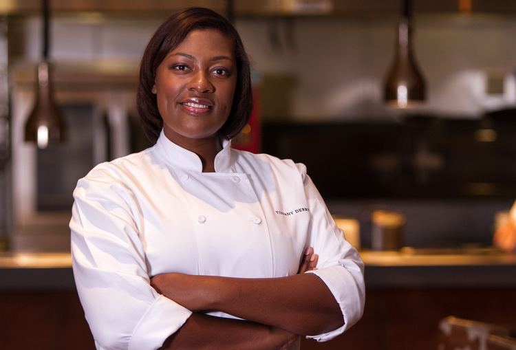 Tiffany Derry smiling with crossed arms on her chest while in the kitchen, with short hair and wearing a white chef gown with her name on it.