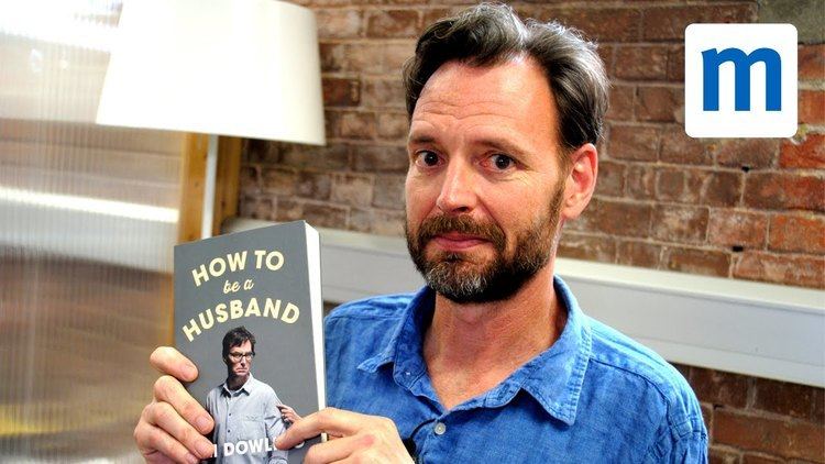 Tim Dowling with a serious face, beard, mustache, and wearing a blue polo shirt while holding a book.