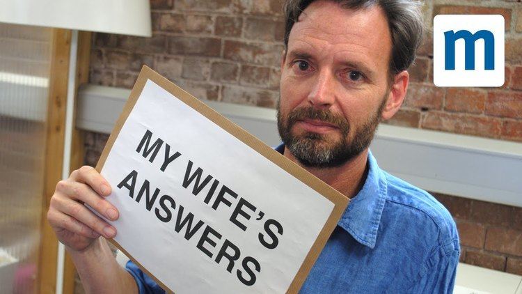Tim Dowling with a serious face, beard, mustache, and wearing a blue polo shirt while holding a paper with a caption, "My Wife's Answers".