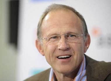 Tim Parks with a big smile while wearing a brown coat, blue and orange striped long sleeves, and eyeglasses