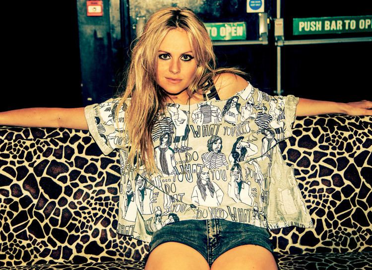Tina O'Brien with a fierce look while sitting on an animal-printed couch, with long blonde hair, wearing a printed shirt and blue shorts.