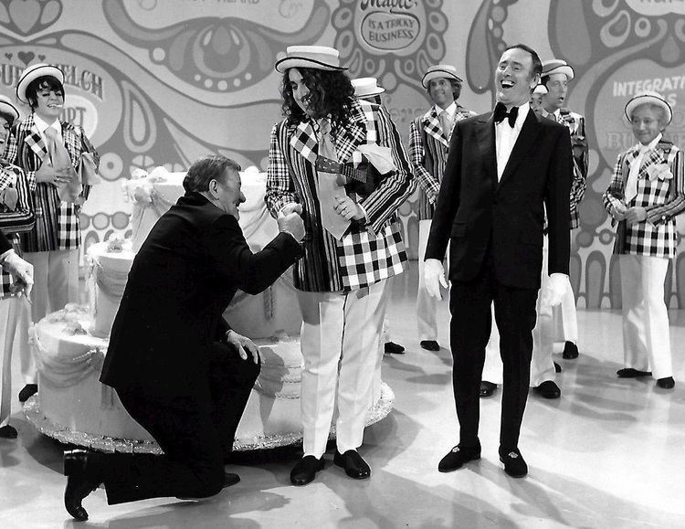 A variety show hosted by Dan Rowan and Dick Martin featuring Tiny Tim wearing a white hat, a checkered suit, white pants, black shoes, and holding a ukelele.