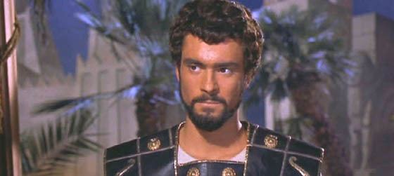 In the movie scene of Jason and the Argonauts 1963 Film, in a room with palm trees and white large walls at the back in front, Todd Armstrong is serious, standing looking to his right, has black hair beard and mustache wearing a white shirt under a black leather armor with round copper plates on.