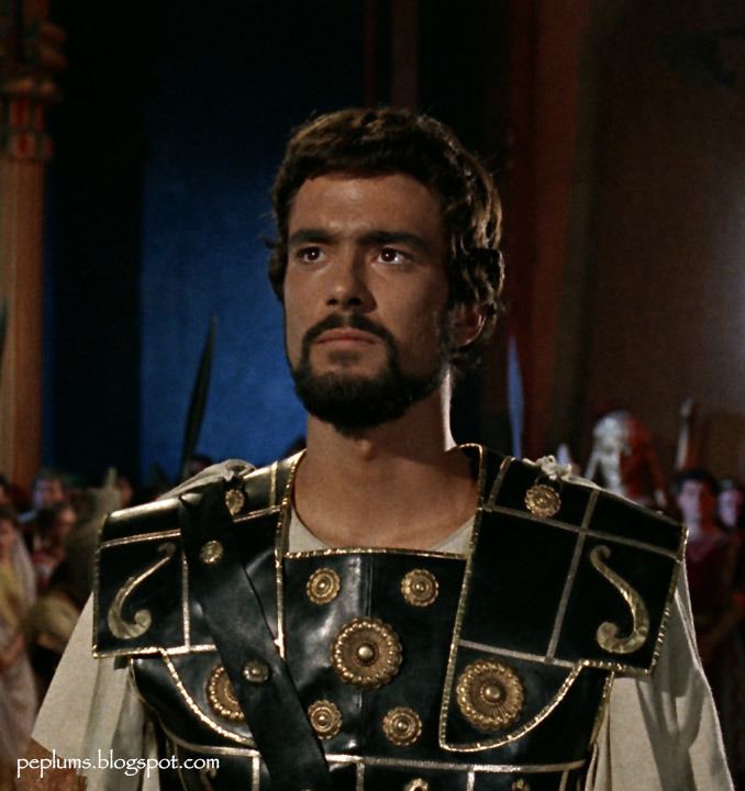 In the movie scene of Jason and the Argonauts 1963 Film, in a dark room with large pillars and men with swords at the back, in front, Todd Armstrong is serious, standing looking up, has black hair beard and a mustache wearing a white shirt under a black leather armor with round copper plates on.