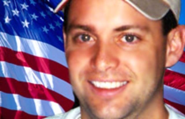 Todd Beamer smiling while wearing a brown cap and white t-shirt and behind him is the flag of the United States of America