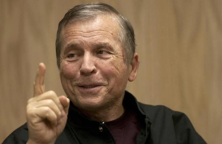 Tom Laughlin smiling while pointing his finger up and wearing a black jacket and maroon shirt