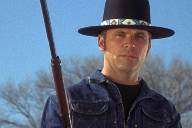 Tom Laughlin holding a rifle while wearing a black hat, t-shirt, and denim jacket in a scene from the 1971 film, Billy Jack