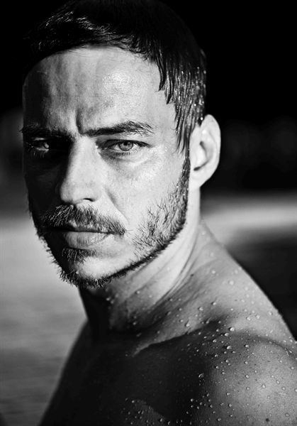 Tom Wlaschiha is serious, has black hair, a beard, and a mustache, his upper body is wet, has a mole on his left arm, topless.