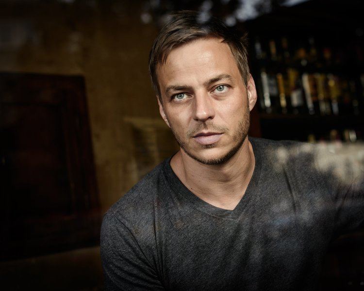 Tom Wlaschiha is serious, has brown hair, a beard, and a mustache, and has a wine cabinet behind (on left) with different kinds of wine, and a door on his right, he is wearing a black sweatshirt.