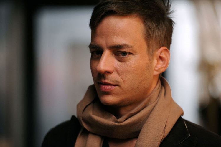Tom Wlaschiha is smiling, and has brown hair, shaved beard, and a mustache, wearing a black suit and a brown scarf.