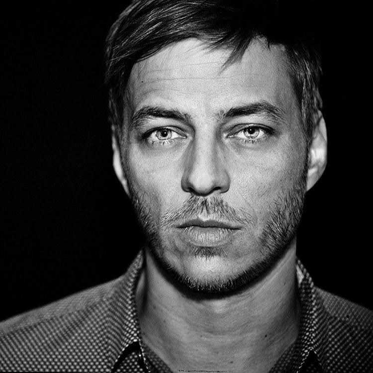 Tom Wlaschiha is serious, has black hair, a beard, and a mustache, with moles on his neck, wearing a black and white checkered polo.